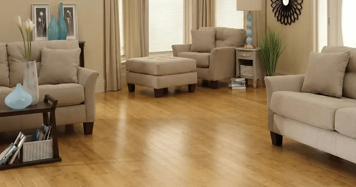 A living room with professionally installed bamboo flooring, beige sofas, a matching ottoman, a side table with a blue lamp, vases, framed pictures, and large windows with blinds and curtains in Madison, WI